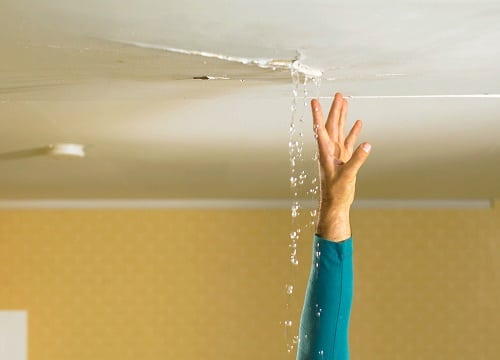 Someone extending their hand up to a leaking ceiling with water from a roof leak