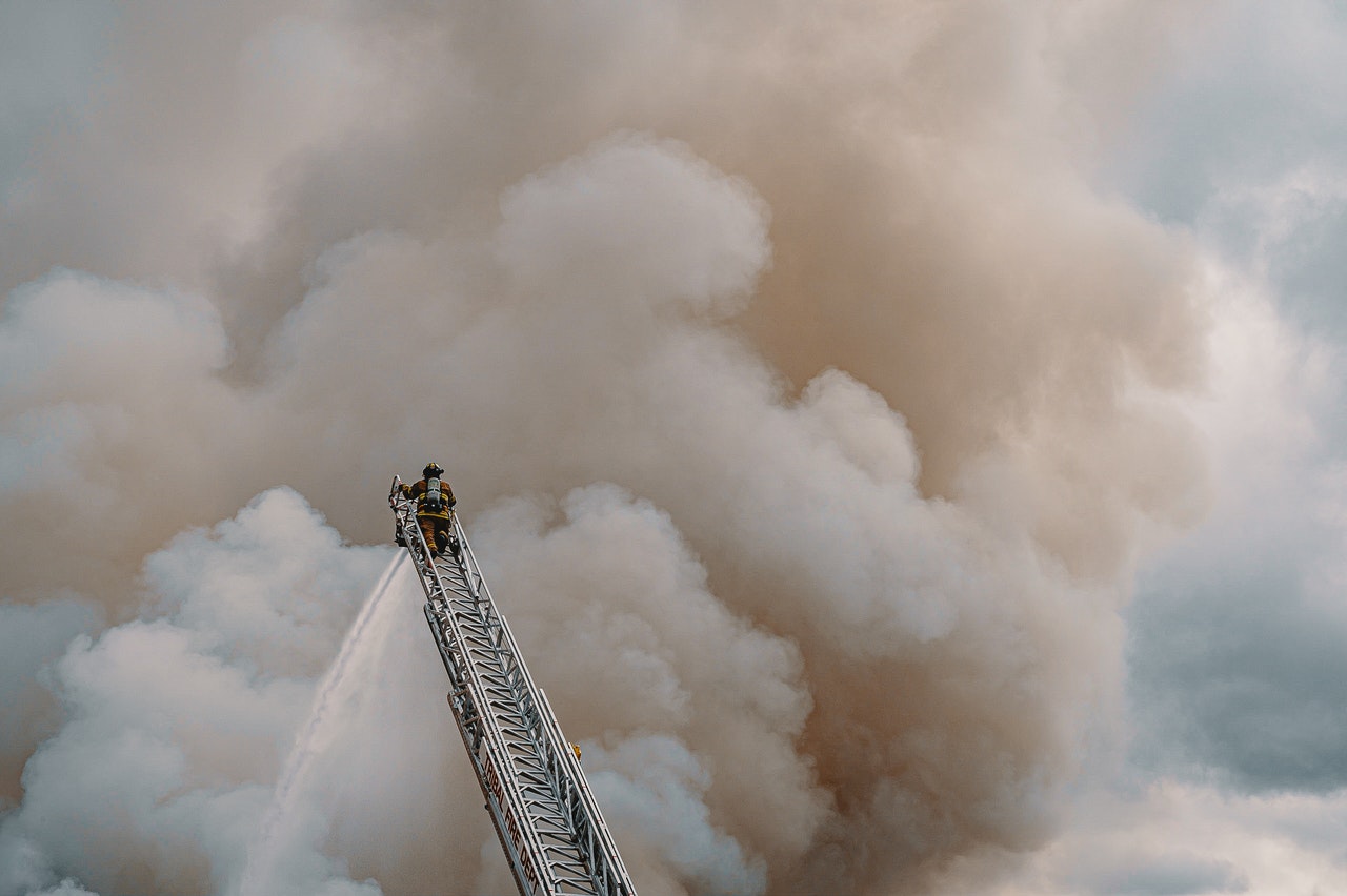 Firefighter at the top of a ladder, spraying water on a fire, with a large billow of smoke in front of him